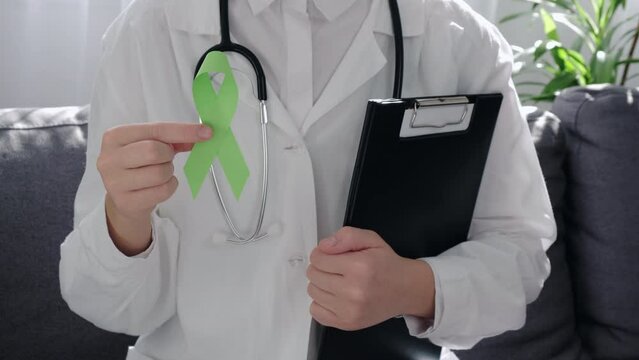 Close-up of female doctor in white coat with stethoscope holding Lime Green Ribbon and clipboard sitting on couch, Mental health awareness and organ donation, world mental health day, world kidney day