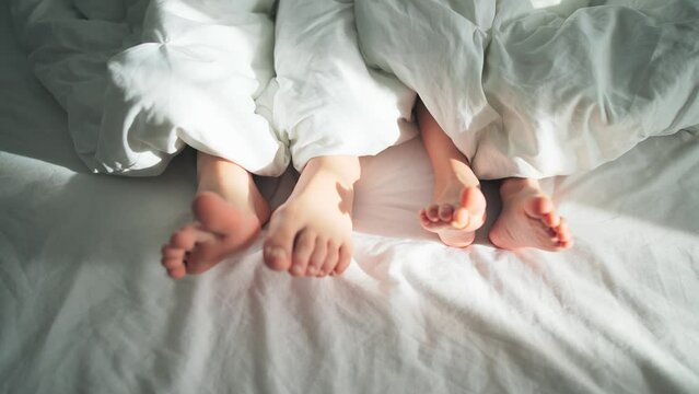 Close-up of bare feet of mother and child from under white blanket on bedding. Parent and newborn lie in bed. Love and care in happy family. New life and parenthood. Sun rays at living room indoors.