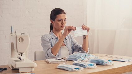Brunette seamstress holding medical mask near sewing machine in atelier.