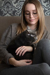 young beautiful woman in glasses at home on the couch holds her gray cat in her arms with a stern character and bright yellow eyes
