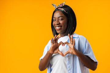 Young african woman smiling and showing a heart shape with hands over yellow background