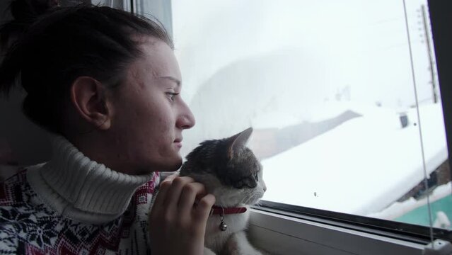 Teen girl with Christmas sweather on the window with her cure cat, petting the cat and looking outside the snowy winter day