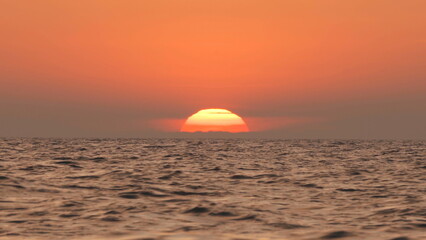 Fototapeta na wymiar A fire red sunset with the sun behind the clouds falling into the sea by an island in the ocean