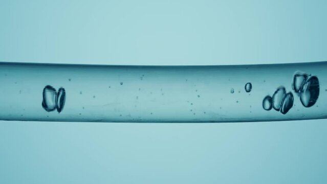 Macro shot of different sized clear bubbles flowing in glass tube with clear liquid on pale blue background. Vessel with clear liquid substance and moving bubbles.