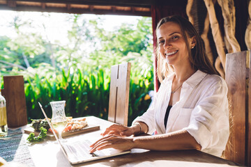 Portrait of joyful female freelancer smiling at camera during time for doing distance startup project on modern laptop technology, joyful digital nomad with netbook laughing while working remotely
