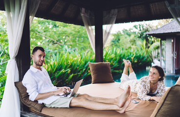Portrait of Caucaisan freelancer spending weekend time with cheerful wife lying at comfortable terrace bed, happy couple in love enjoying digital nomad lifestyle using laptop for remote working