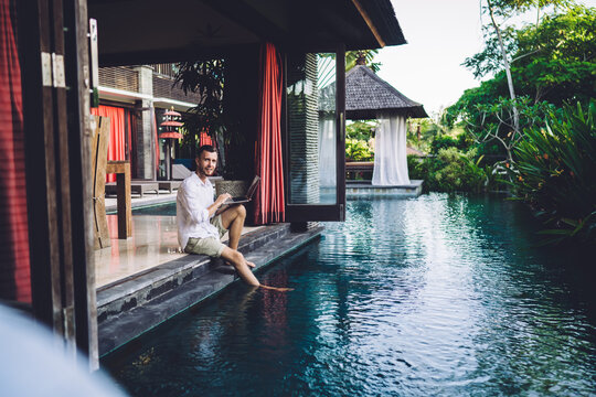 Portrait of Caucaisan digital nomad looking at camera while doing distance job at pool terrace of resort villa, skilled male freelancer with modern laptop technology posing at Indonesian courtyard