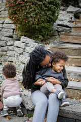 Biracial mom cuddling laughing toddler son on steps in backyard in autumn