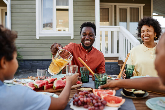 Portrait of smiling black man pouring orange juice to glass during family gathering outdoors - Powered by Adobe