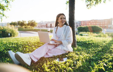 Smiling woman sitting in green lawn and using laptop in park
