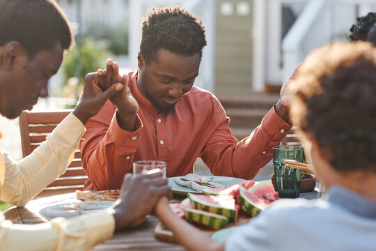 Portrait of young black man saying grace at table outdoors during family gathering and holding hands in sunlight