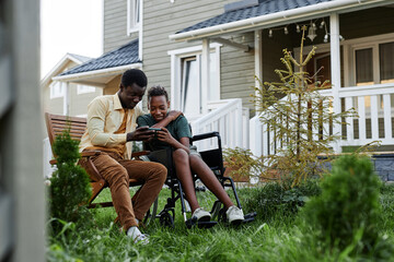Full length portrait of smiling teenage boy looking at smartphone screen while spending time with father outdoors by house