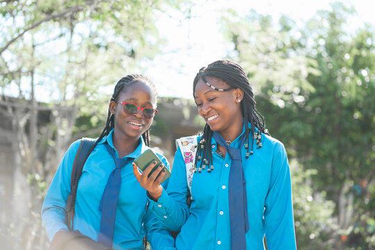 Due giovane studentesse africane in uniforme scolastica guardano contenuti divertenti sullo smartphone mentre vanno a scuola.