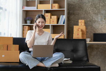 Asian woman sitting on sofa at home working freelance using laptop with box, successful Asian woman with raised hand, online marketing packaging and delivery box, SME concept