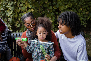 Young Black girl blowing bubbles with her family on bench