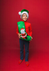 a boy in a knitted red sweater and a Santa Claus hat holds a Christmas boot for gifts. Cute baby is waiting for Christmas and a new kind. insulation on a red background.