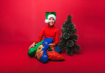 a little boy wearing a Santa Claus hat is sitting on the floor with a small Christmas tree and New Year's gifts on a red background with a space for text. the concept of Christmas