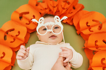 Halloween portrait of adorable baby lying on green background next to pumpkin garland, holding...
