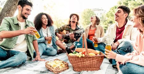 Group of happy friends having fun at picnic playing guitar and singing at sunset - Youth friendship concept with boys and girls drinking beer at the bar-b-q party.