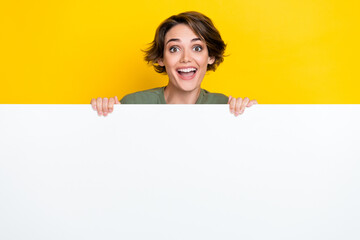 Portrait of impressed girl bob hairstyle wear khaki t-shirt hold white banner open mouth speechless isolated on yellow color background