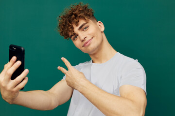 a joyful, cute man takes a selfie on his smartphone showing a peace sign with his fingers, standing on a green background in a gray T-shirt