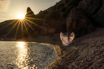 A mysterious female silhouette with long braids stands on the sea beach with mountain views, Sunset rays shine on a woman. Throws up a long white dress, a divine sunset.