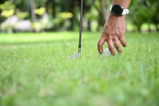Golfer's hand placing a white golf ball on a embroidered tee on a green lawn with blurred green golf course background. The left hand is not gloved, placing or removing the golf ball on the tee.
