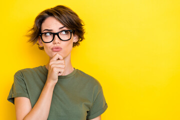 Portrait of thoughtful woman with bob hairdo dressed khaki t-shirt hand on chin look empty space isolated on yellow color background