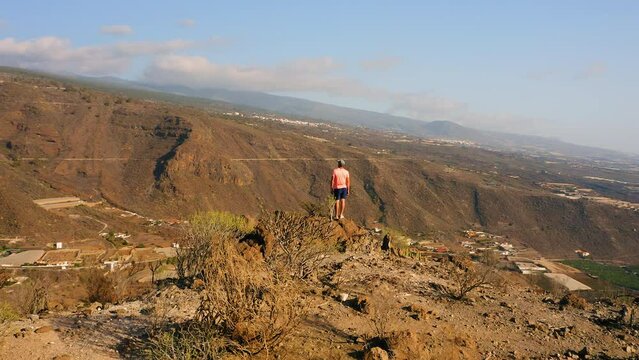 Tenerife Canary Island, mountain landscape. Tourist man at the edge of the sheer cliffs. Hiking area. Amazing nature, scenery. Village Tamaimo in a gorge. Drone circular flight around a person.