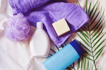 Purple towel, sponge, white shampoo bottle, blue shower gel packaging, soap bar and palm leaves. Flat lay photography, top view