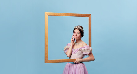Emotional girl, actress wearing medieval princess or countess dress standing with picture frame over blue background. Portrait through picture frame.