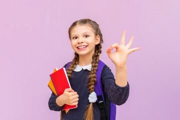 A little schoolgirl with pigtails shows an ok sign with her hand and smiles broadly on a pink isolated background. Advertising of educational courses for schoolchildren.