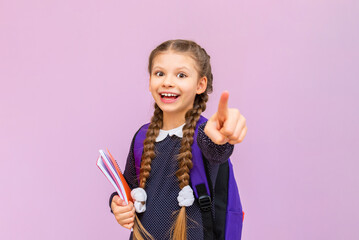 A little schoolgirl with pigtails points her index finger forward and smiles on a pink isolated background. Advertising of educational courses for schoolchildren.