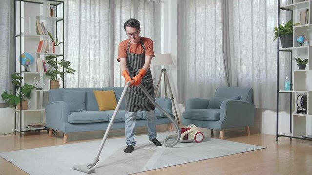 Asian Male Housekeeper With An Apron Enjoys Vacuuming The Floor At Home
