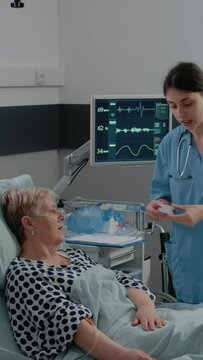 Vertical Video: Woman Working As Nurse Using Oximeter For Oxygen Saturation Measurement To Cure Elder Patient In Bed. Medical Assistant Putting Tool On Hand Of Woman With Oxygen Tube And IV Drip Bag