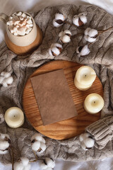 Cozy home flat lay: a cup of hot chocolate or cocoa with marshmallows on a wooden board, a wool sweater, candles and a sprig of cotton. Top view. Place for text.