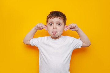 A little caucasian boy in a white t-shirt shows tongue pulling the ears. The child grimacing and showing a funny face isolated on a color yellow background