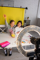 Naklejka na ściany i meble smiling girl showing spiral toy while sitting near friend and blurred digital camera in circle lamp