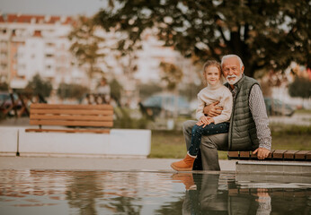 Grandfather spending time with his granddaughter by small water pool in park on autumn day