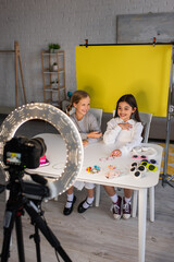 Fototapeta na wymiar smiling preteen video bloggers talking near different accessories and digital camera in ring light on yellow background at home