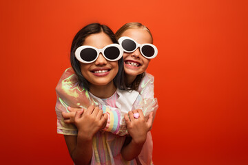 happy girls in stylish sunglasses hugging and smiling isolated on orange