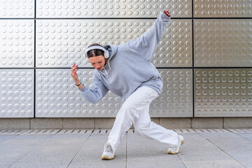 Young woman with headphones listening to urban music and dressed in white pants and gray hoodie dancing in front of metallic background urban dance