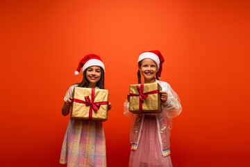 happy friends in santa hats smiling at camera while holding gift boxes isolated on orange
