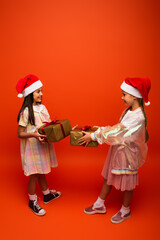 full length of smiling preteen girls in santa hats holding christmas presents on orange background