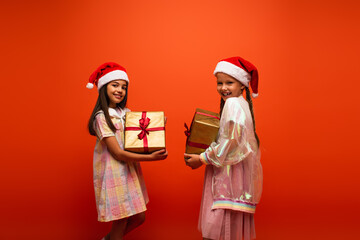 cheerful girls in santa hats holding gift boxes and smiling at camera isolated on orange