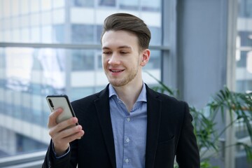 Fototapeta premium Portrait of happy handsome guy, young European blonde man in shirt is using his cell mobile phone, looking at screen of smartphone, reading or typing message and smiling in office building indoors.