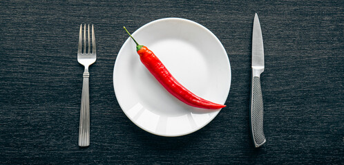 Flat lay, chili peppers on a white plate and cutlery on a black background.