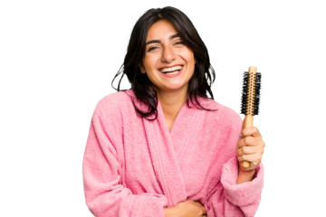Young Indian woman in a bathrobe holding an hairbrush isolated laughing and having fun.