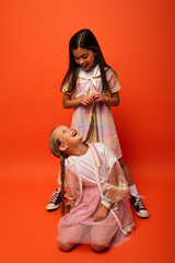 Fototapeta premium brunette girl holding pigtail of cheerful friend sitting on orange background