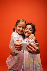 joyful preteen girls in trendy clothes embracing while taking selfie on smartphone isolated on orange